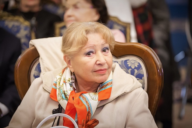 Rodica Popescu Bitănescu, stând pe scaun, îmbrăcată intr-un palton bej și o eșarfă colorată