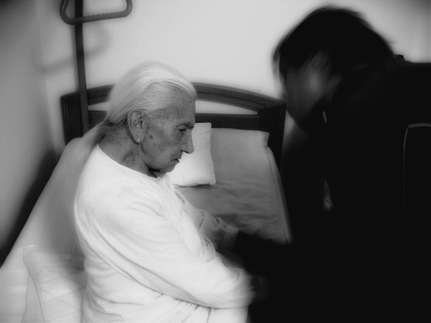 femeie cu demență care stă pe un pat și este supravegheată de cineva