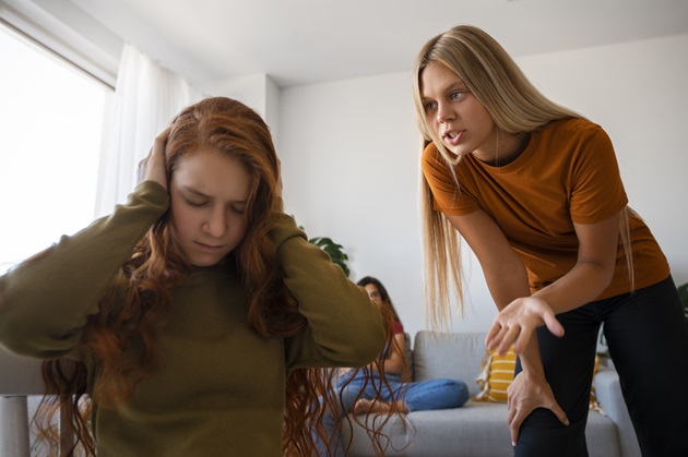 O femeie blondă care stă în picioare și țipă la alta roșcată care își astupă urechile cu mâinile