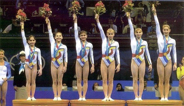 Simona Amânar ridicând un buchet de flori în mână, pe un podium olimpic alături de alte tinere gimnaste