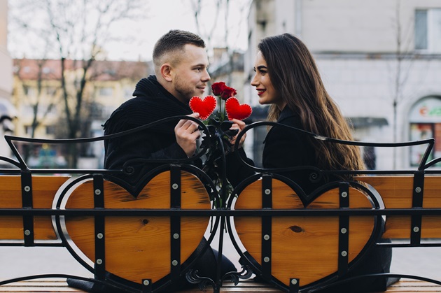cuplu frumos pe o bancă romantică tinând în mână niște inimioare roșii și niște flori