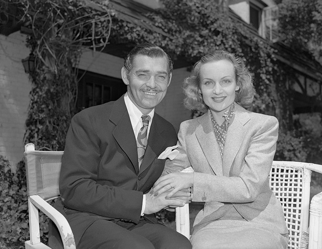 Poză cu Clark Gable și Carole Lombard ținându-se de mână, în timp ce stau așezați pe o bancă 