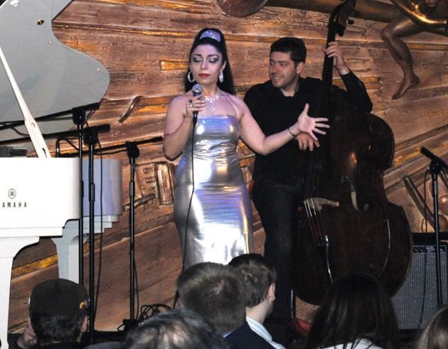 Anca Parghel pe o scena în fața unui public de tineri, alături de un coleg, purtând o rochie elegantă, argintie