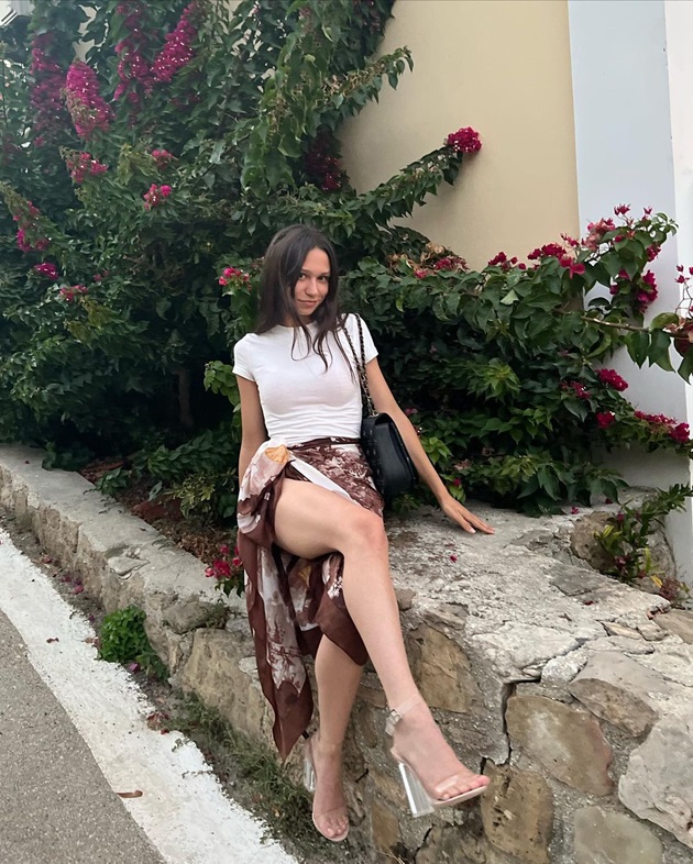 Maia Trandafir îmbrăcată într-o fustă maronie, cu piciorul la vedere, un tricou alb și sandale cu toc