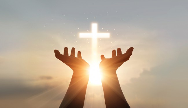 doua maini care se roaga spre cer cu cruce luminata in fundal