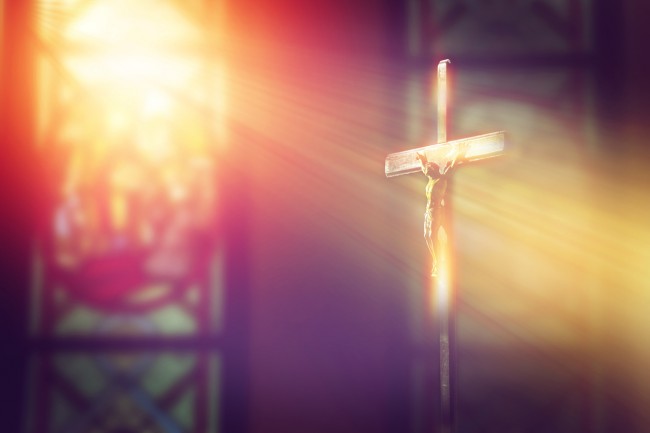 imagine din biserica cu raze de soare trecand prin vitralii luminand o cruce cu Iisus Hristos