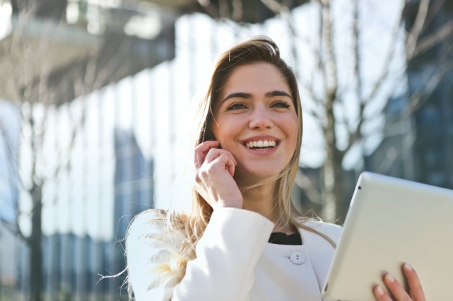 femeie blonda si frumoasa imbracata in palton alb cu tableta in mana care vorbeste la telefon, femeie de afaceri cu succes