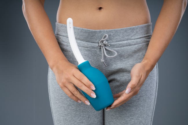 femeie imbracata in pantaloni de trening gri care se pregateste sa faca un dus vaginal; dispozitiv pentru dus vaginal