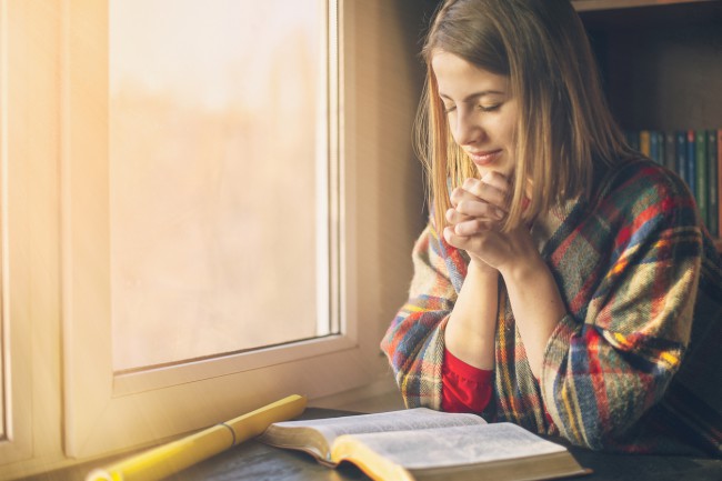 fata care se roaga in fata unui geam cu sal pe umeri si citind dintr-o carte de rugaciuni