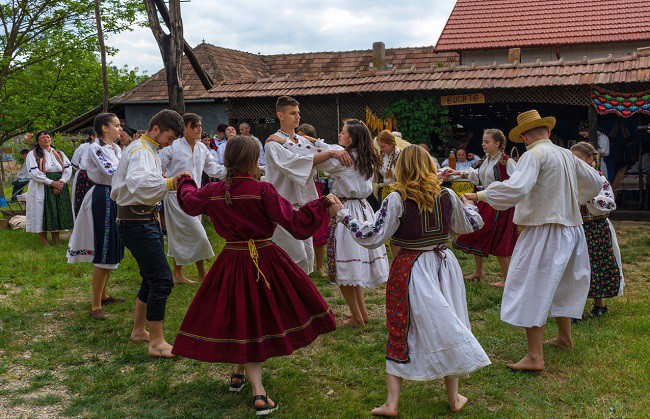 grup de fete si baieti imbracati in costume populare care danseaza in fata unei case in stil traditional