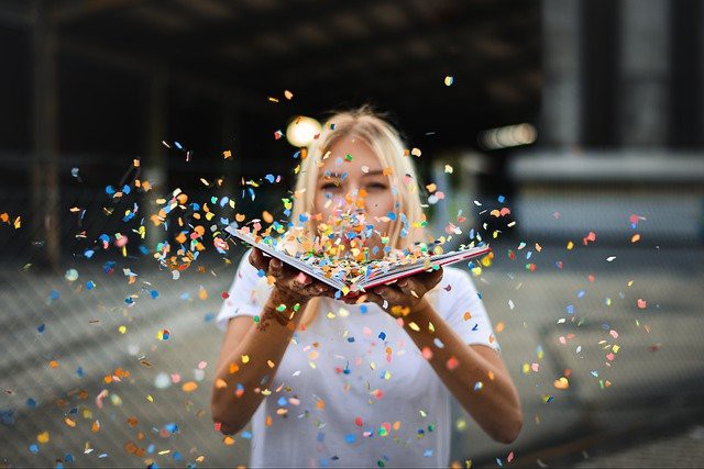 femeie blonda fericita care arunca cu confetti colorat dintr-o carte