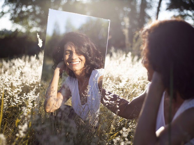 femeie care se priveste in oglinda intr-un camp plin cu flori