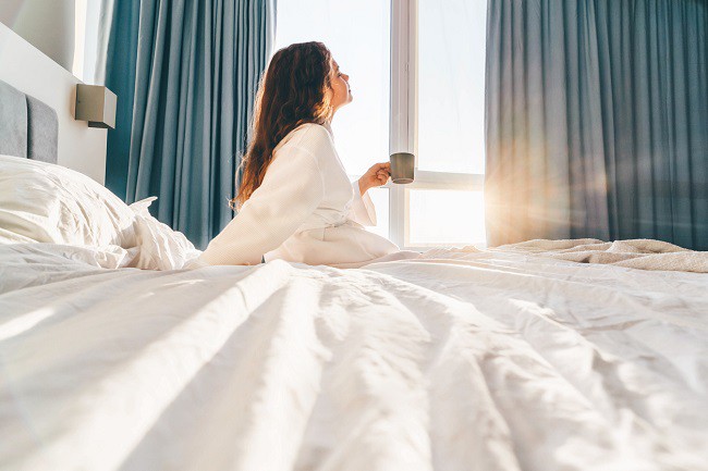 fata care sta in pat si bea cafea privind pe geamul unde rasare soarele