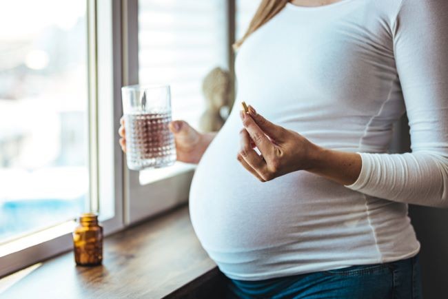 femeie insarcinata care sta langa geam si ia vitamine cu un pahar cu apa