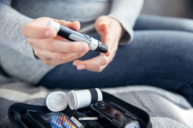 femeie imbracata in blugi care isi monitorizeaza glicemia cu un glucometru aflat pe masa langa ea