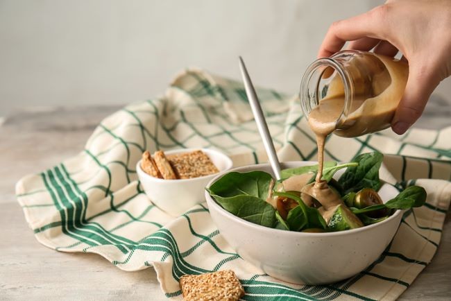 imagine cu bol de salata pe un prosop de bucatarie in care o mana adauga pasta de tahini 