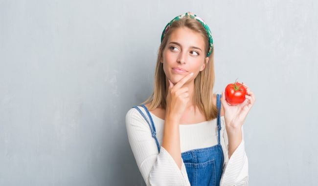 fata imbracata intr-o salopeta de blugi care sta pe fundalul unui perete si se gandeste daca sa manance o rosie