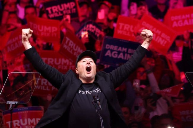 elon musk in costum ridica mainile in sus intr-o sala cu oameni cu mesaje pro trump