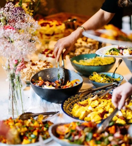 masa de eveniment decorata cu flori de pe care diverse persoane se servesc cu mancare