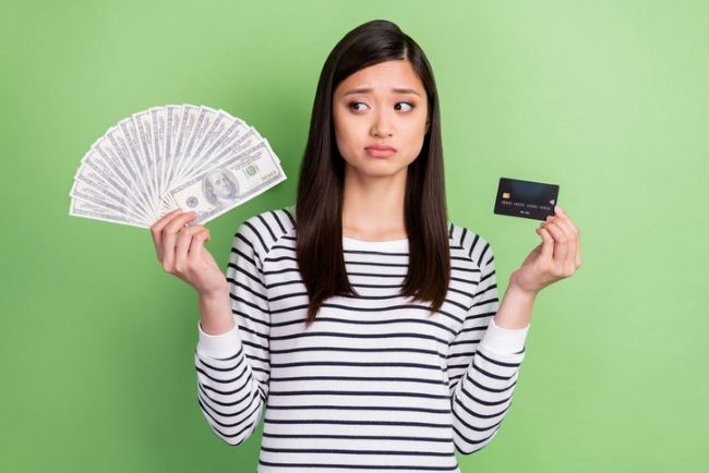 femeie asiatica in bluza in dungi care e preocupata si tine intr-o mana bani iar in cealalta un card