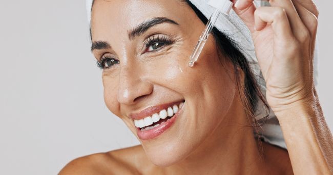 femeie de varsta mijlocie, bruneta si cu prosop in cap, care aplica un serum