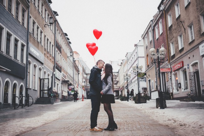 O fată și un băiat care se sărută și baloane în formă de inimă deasupra lor