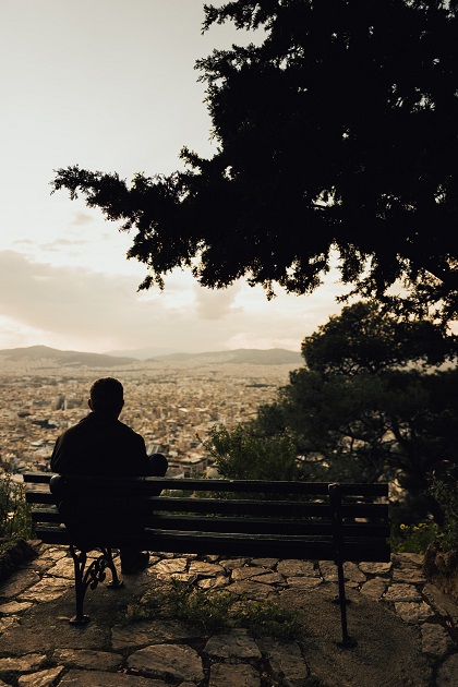 Silueta unui bărbat care stă pe o bancă așezată pe un deal de lângă un orășel.