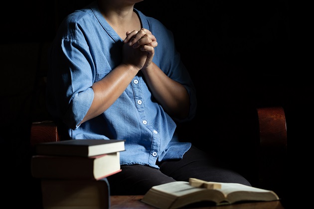 Femeie care se roagă într-o încăpere întunecată înconjurată de cărți de rugăciune