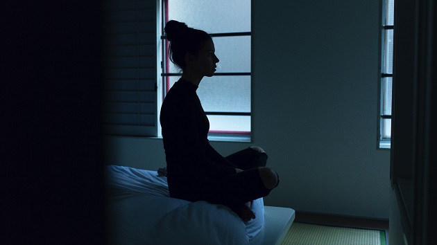 Femeie care stă îngândurată la marginea patului într-o cameră semiîntunecată în timp ce se uită în gol. 