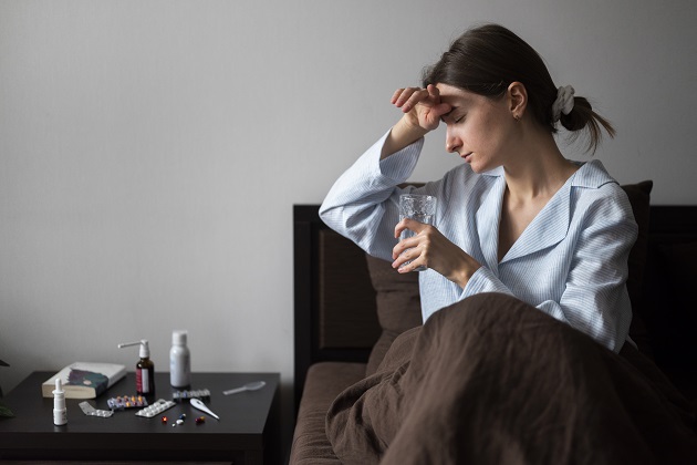 Femeie bolnavă stând în pat lângă o noptieră plină de medicamente, vizibil afectată de răceală. 