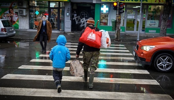 barbat cu sacosa in spate si copil de mana traverseaza trecerea de pietoni pe ploaie printre masin