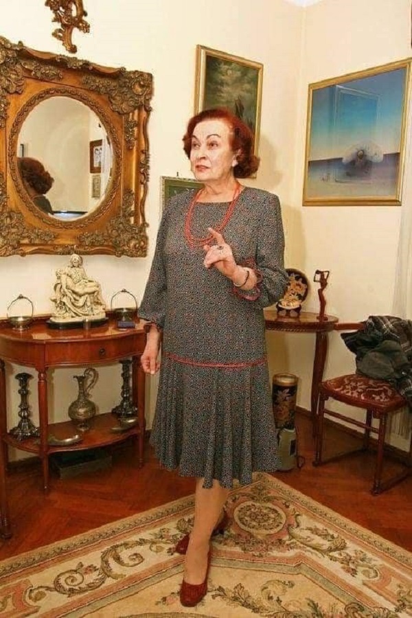 sanda taranu imbracata in rochie neagra cu buline albe sta in picioare intr-un living