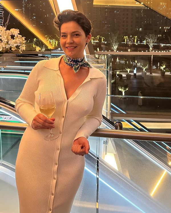 maria dinulescu cu rochie alba si un pahar de vin alb in mana pe un balcon