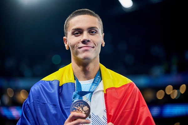 david popovici cu steagul romaniei in spate tine medalia de aur in mana