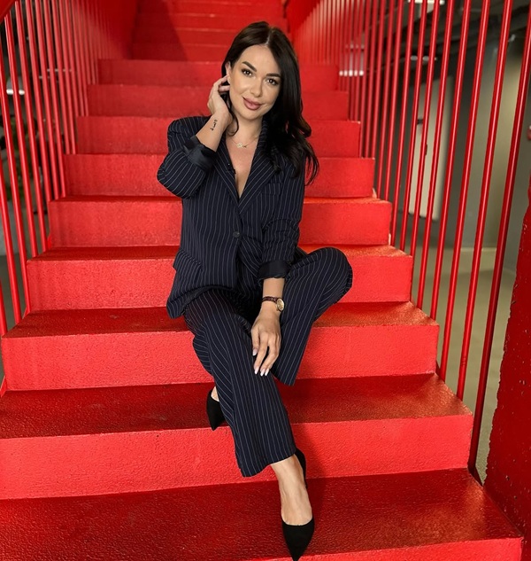 deea maxer in costum cu pantaloni negru sta pe scari de culoare rosie si isi trece mana prin par