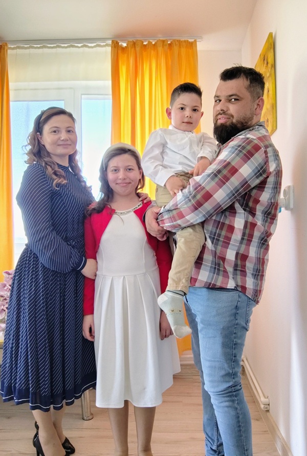 femeie imbracata in rochie albastra midi alaturi de o fetita in rochie alba si un barbat in camasa cu carouri ce tine un copil in brate intr-o sala de clasa