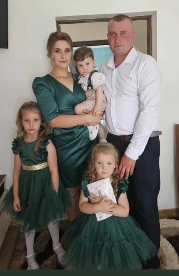 un barbat in camasa alba alaturi de o femeie in rochie verde care tine un copil in brate si doua fete