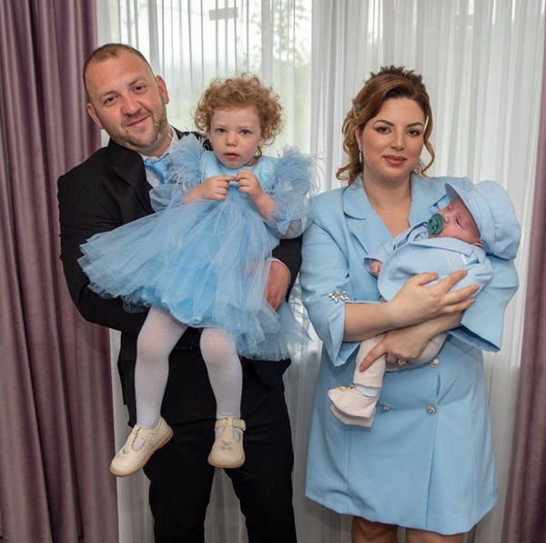 mama imbracata in rochie albastra isi tine bebelusul in brate alaturi de sot imbracat in negru care isi tine fetita in rochie bleu