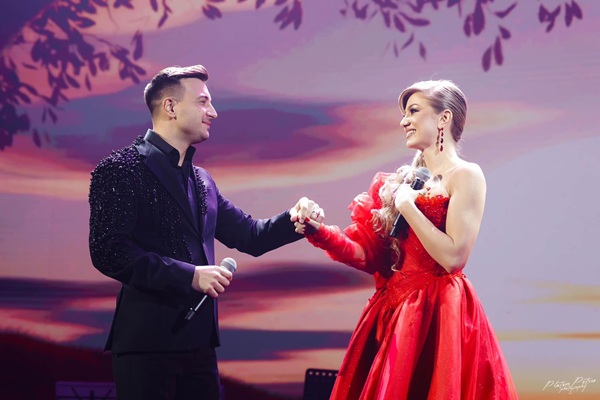 valentin sanfira imbracat in costum negru cu microfon in mana pe scena cu sotia lui imbracata in rochie lunga rosie eleganta