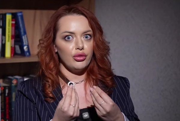 femeie roscata cu buzele rosii in costum negru explica ceva gesticuland din maini