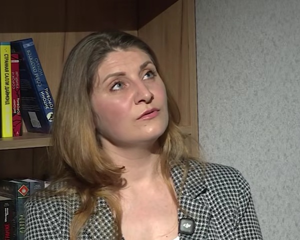 femeie blonda cu sacou gri langa o biblioteca priveste in sus