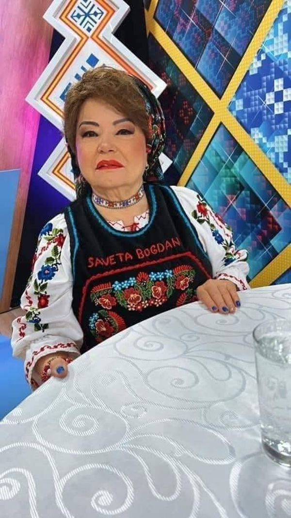 saveta bogdan in costum popular sta la masa cu un pahar de apa in fata