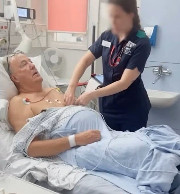 jeremy clarkson pe un pat de spital invelit cu o patura langa o asistenta imbracata in albastru