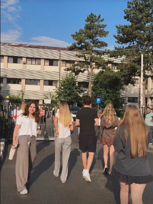 fiica ancai serea in pantaloni gri si tricou alb intra pe poarta liceului alaturi de alti colegi