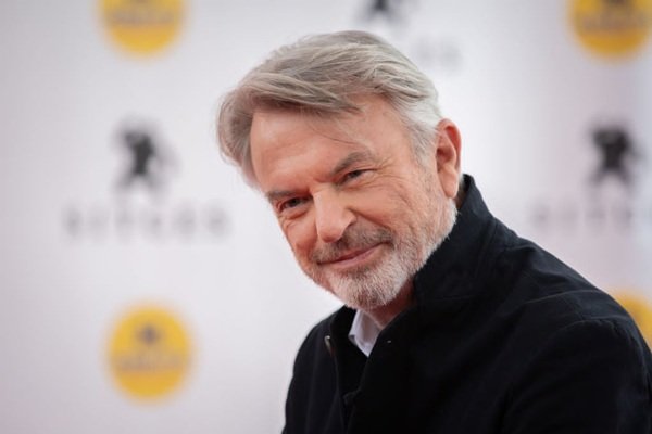 actorul sam neill cu parul grizonat in camasa neagra priveste spre camera