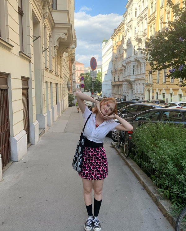 fata cu ochelari de vedere parul blond tuns scurt imbracata in bluza alba si fusta rosie in carouri pozeaza pe strda