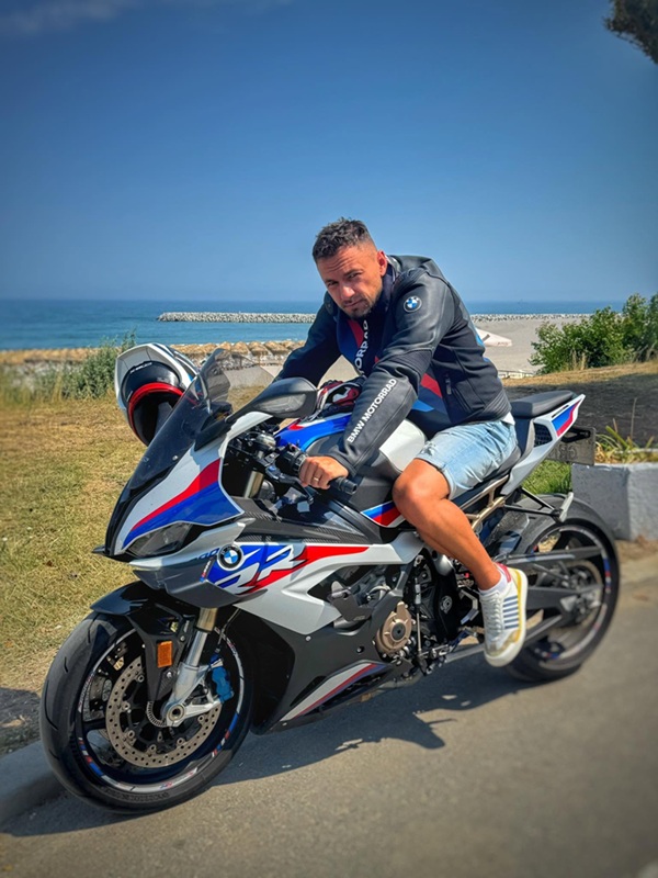 un barbat imbracat in negru si pantaloni trei sferturi pe o motocicleta
