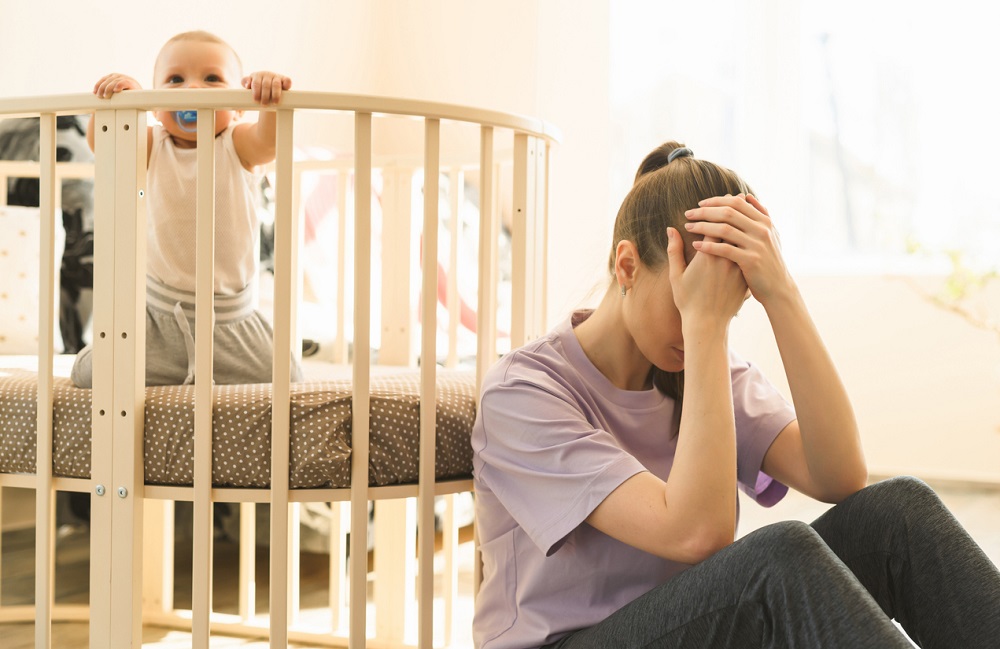 Copil mic in patut, cu o mama epuizata, cu mainile tinandu-se de cap, asezata la picioarele patului