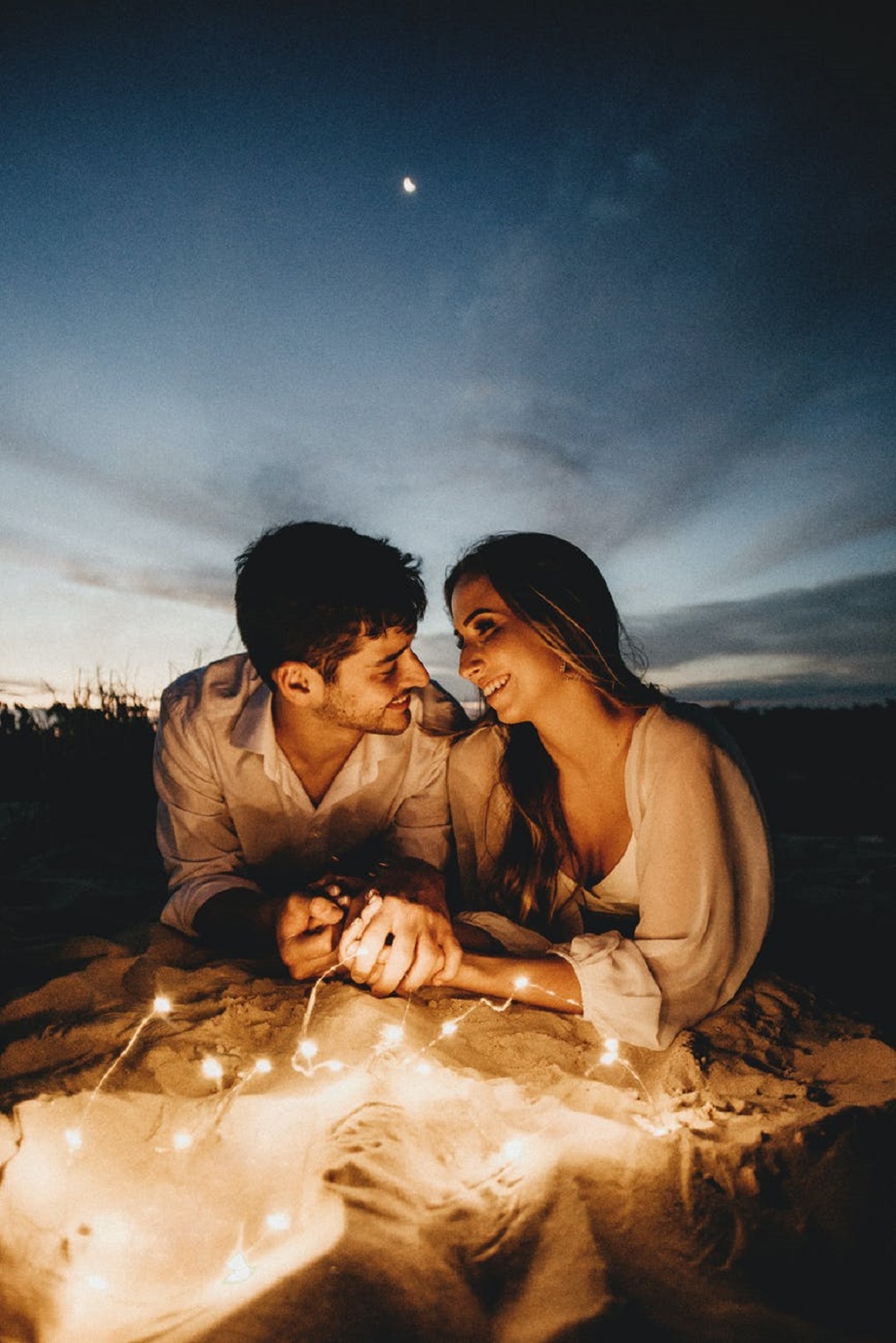 Îndrăgostiți care se țin de mână și se bucură de o întâlnire în aer liber, noaptea, într-o atmosferă romantică, înconjurați de beculețe și cu luna în fundal