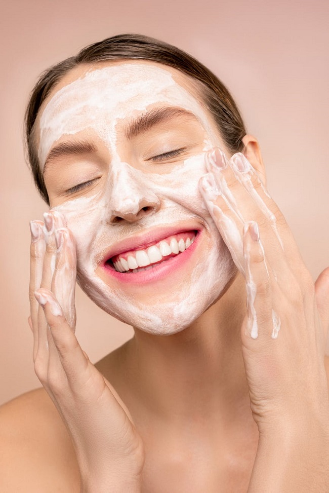 Femeie zâmbitoare care își aplică spuma de curățare pe față, în timpul rutinei de îngrijire.